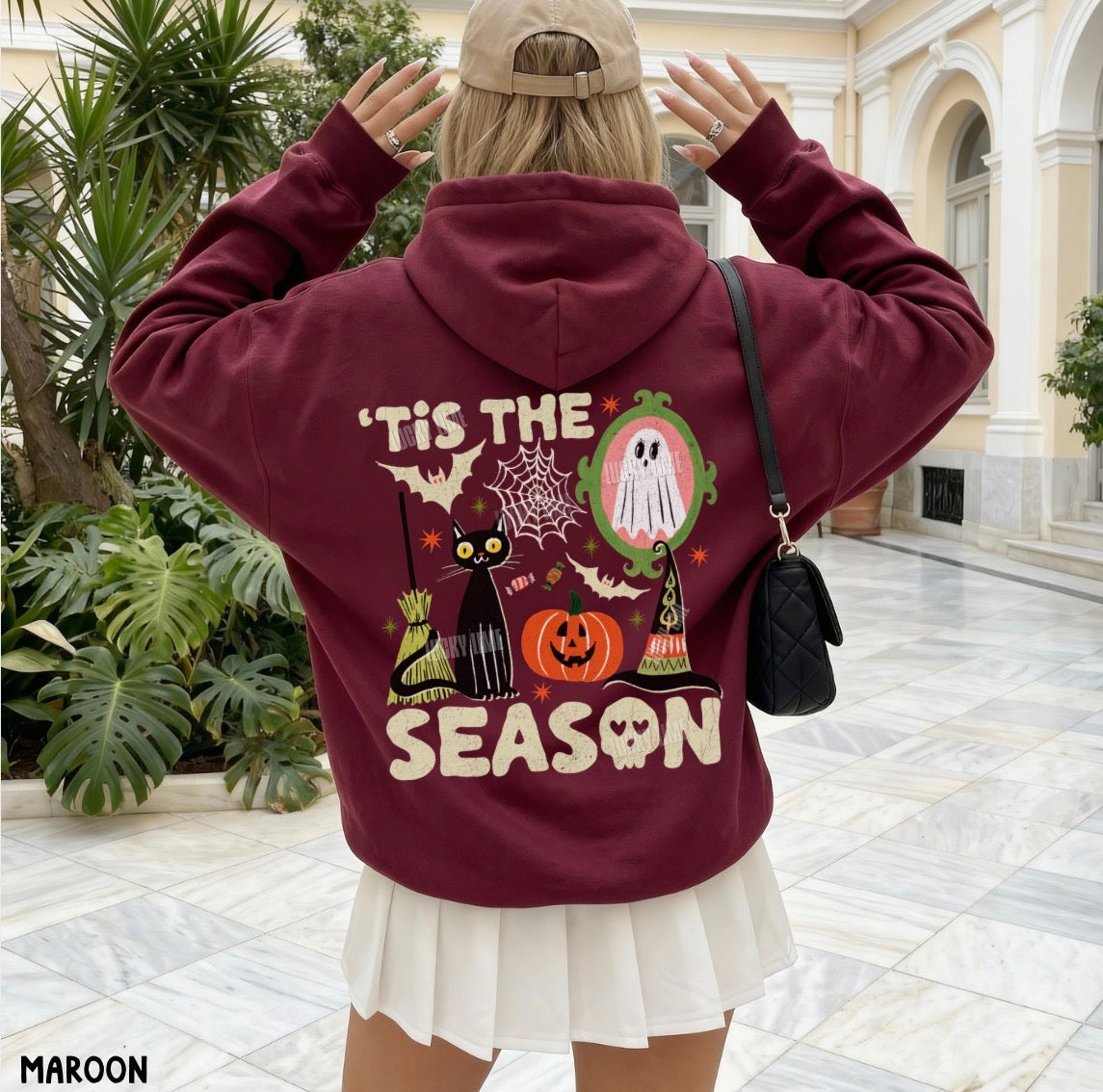 A woman viewed from behind wearing a maroon hoodie with a large Halloween-themed graphic on the back, including a ghost, pumpkins, bats, and the text “'TIS THE SEASON.” She is standing in a tiled indoor courtyard with plants and a building facade in the background.