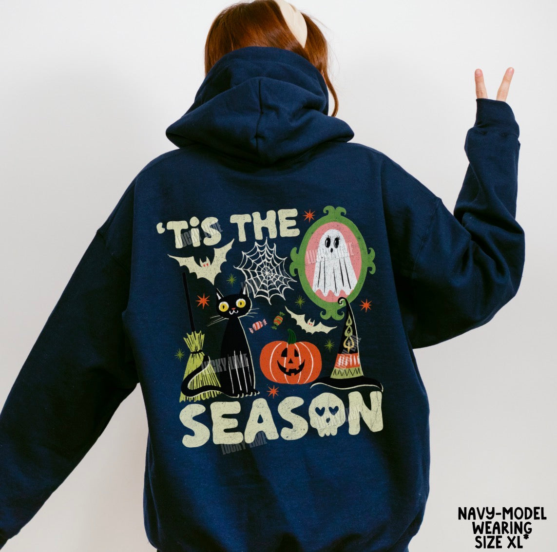 A person shown from the back wearing a navy hoodie with a large Halloween-themed graphic on the back, including a ghost, pumpkin, cat, spiderwebs, bats, and the text “'Tis the Season.”.