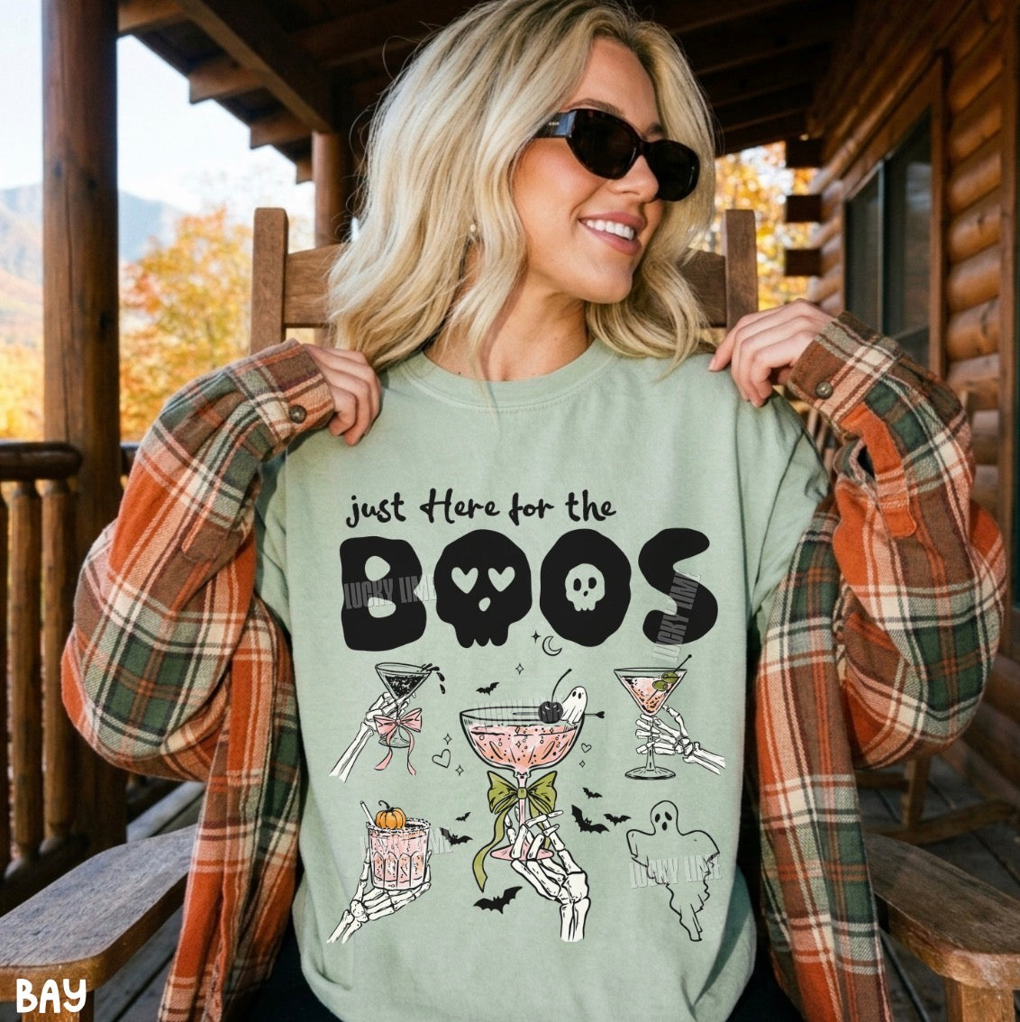 A woman wearing a sage green graphic t-shirt with a Halloween design, styled with an orange and green plaid flannel shirt, sitting on a wooden porch.