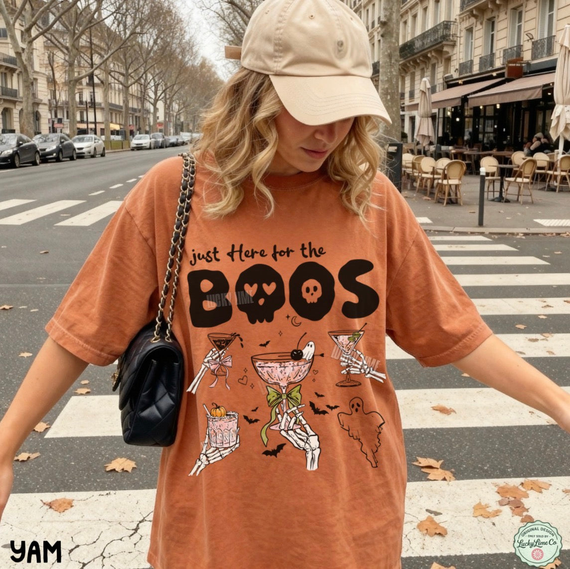 A woman wearing an oversized burnt orange graphic t-shirt with a Halloween theme, a beige baseball cap, and a black shoulder bag, standing on a city street crosswalk.