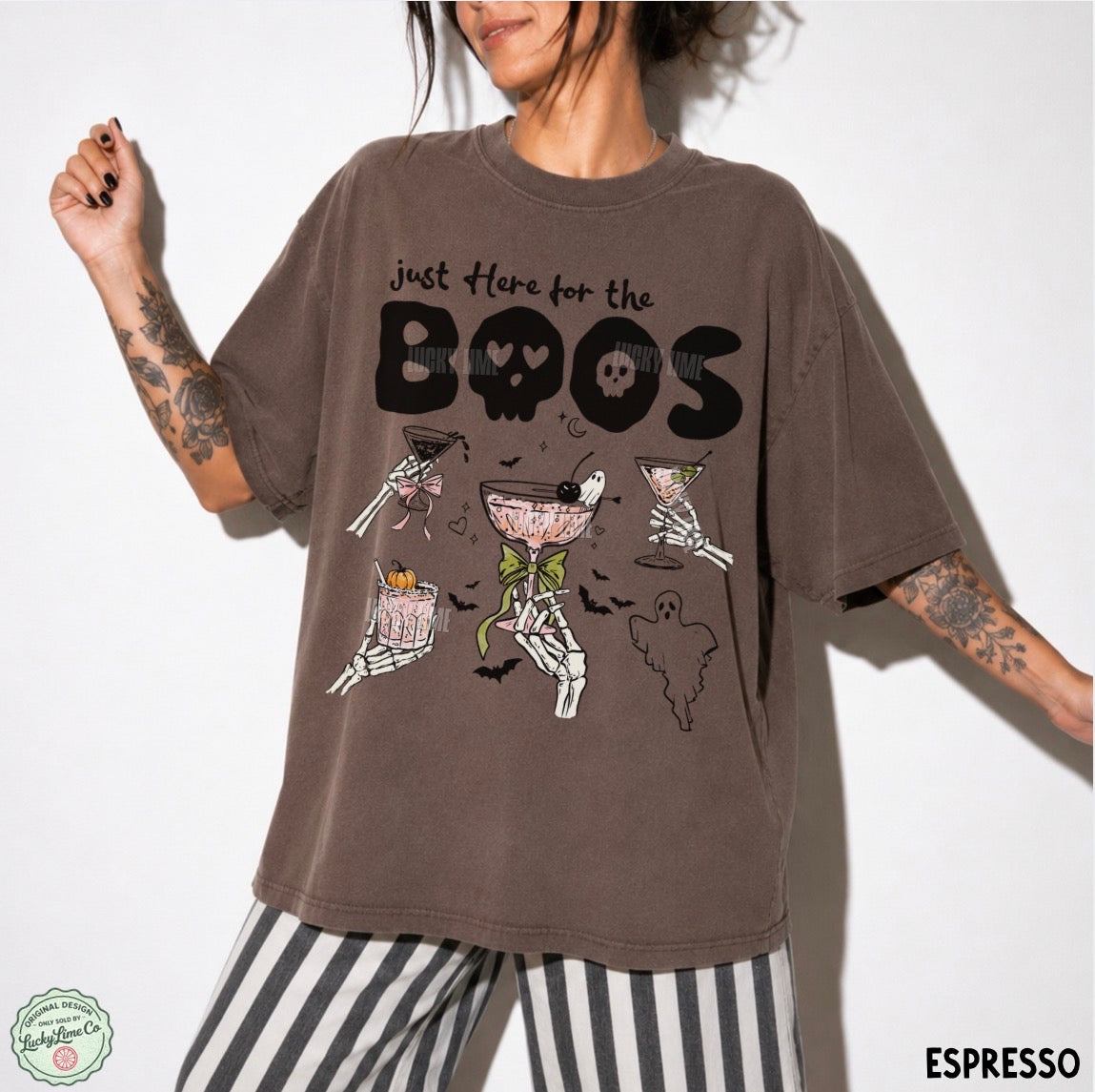 A woman wearing an oversized brown graphic t-shirt with a Halloween-themed design featuring skeleton martinis and the text 'just here for the BOOS'. She is also wearing black and white striped pants, photographed against a plain white background.