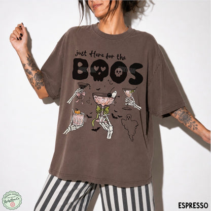 A woman wearing an oversized brown graphic t-shirt with a Halloween-themed design featuring skeleton martinis and the text 'just here for the BOOS'. She is also wearing black and white striped pants, photographed against a plain white background.