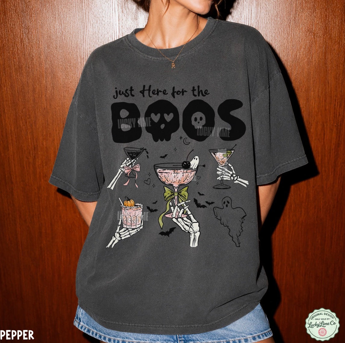 A person wearing an oversized charcoal gray graphic t-shirt with a Halloween theme, featuring skeleton hands holding cocktails and the text 'just Here for the BOOS' with skulls. The t-shirt is shown against a dark wood-paneled background.