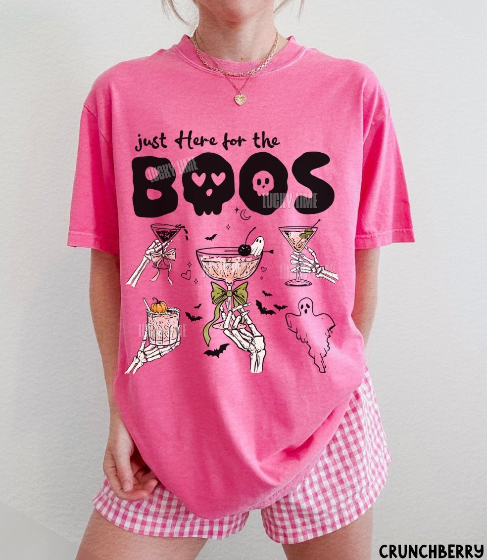 A person wearing a bright pink graphic T-shirt with the text 'just Here for the BOOS' and Halloween-themed illustrations, paired with pink-and-white gingham shorts. The brand name CRUNCHBERRY appears in the bottom right corner.