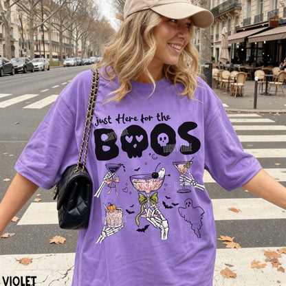 A woman wearing a purple oversized graphic T-shirt with the text 'Just Here for the BOOS' and Halloween-themed illustrations, standing on a city street crosswalk.