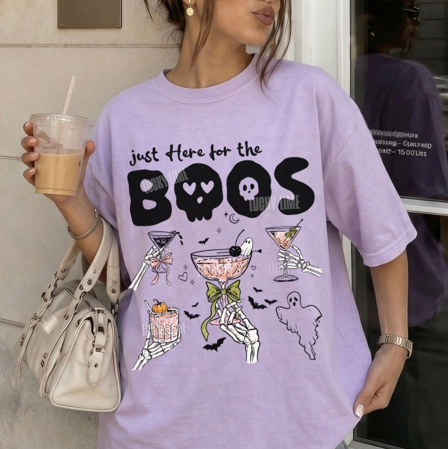 A woman wearing an oversized lavender graphic T-shirt with Halloween-themed illustrations and the text 'just Here for the BOOS'. She is holding an iced coffee and carrying a light beige handbag, standing in front of a building wall.