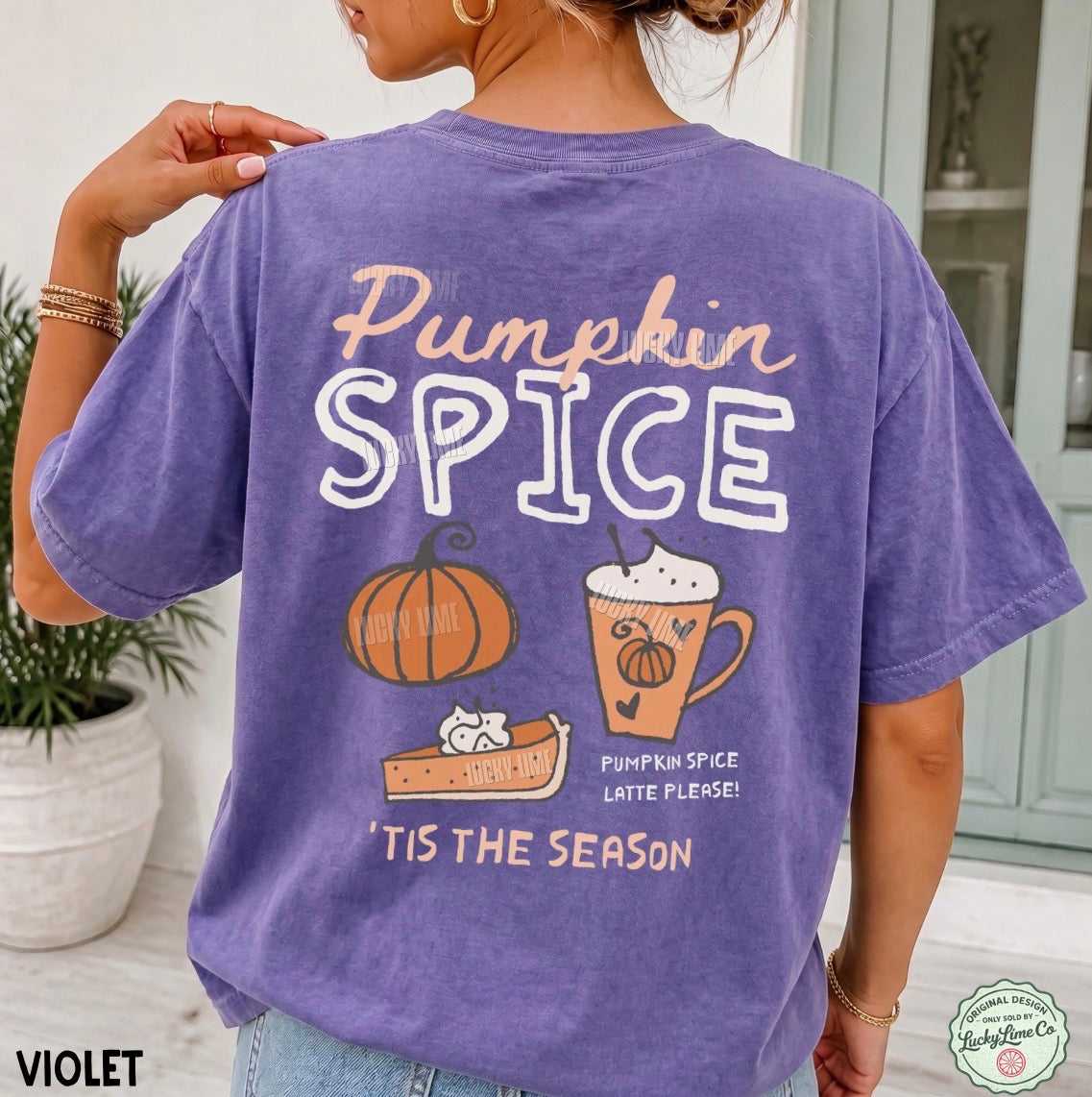A woman wearing a violet short-sleeve graphic t-shirt, viewed from the back. The shirt features a colorful 'Pumpkin Spice' autumn-themed design with a pumpkin, a latte, and a cat.