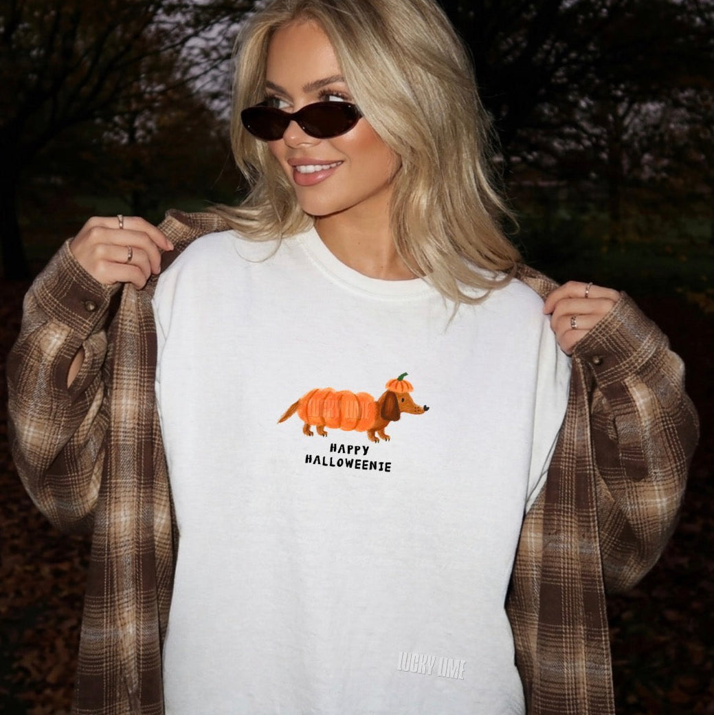 A woman outdoors wearing a white graphic t-shirt with an orange pumpkin-dog design and the text 'HAPPY HALLOWEENIE', holding open a brown plaid flannel shirt. She is wearing dark sunglasses.