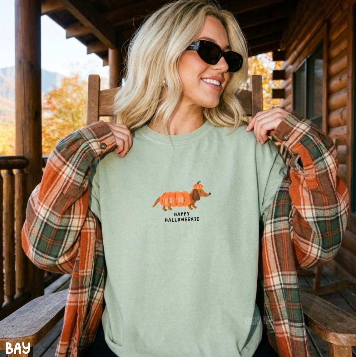 A woman wearing a sage green crewneck sweatshirt with a cartoon orange dachshund and the text 'HAPPY HALLOWEENIE' on the chest, layered under an open rust-orange plaid flannel shirt on a wooden porch.