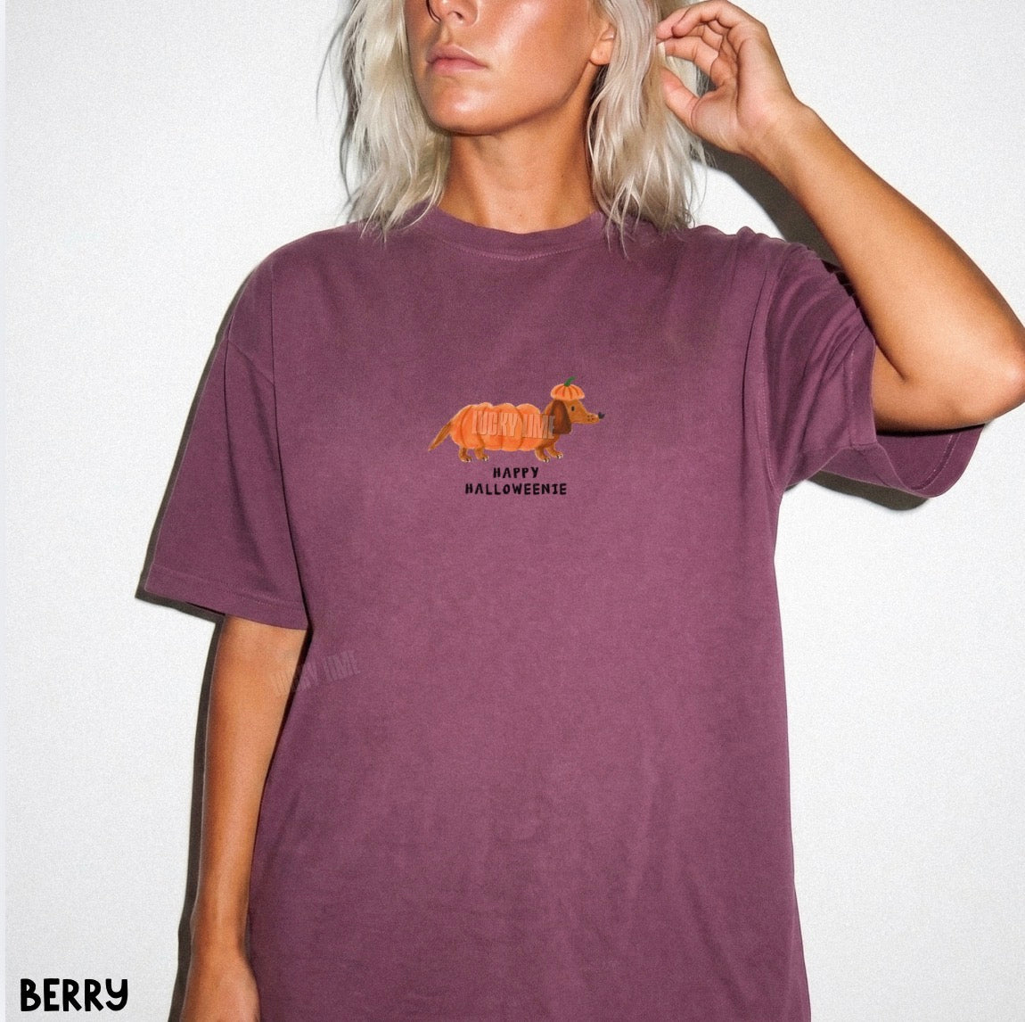 A woman wearing a berry purple short-sleeve t-shirt with a graphic of a dachshund wearing a pumpkin and the text 'HAPPY HALLOWEENIE'.