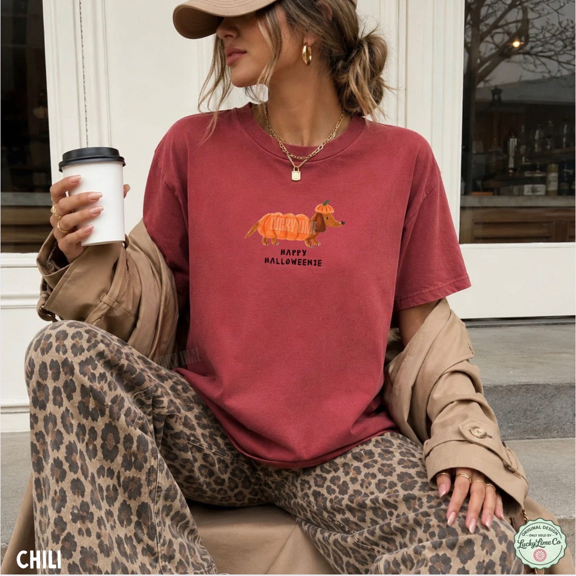 A woman wearing a relaxed-fit dusty rose red graphic t-shirt with a 'Happy Halloween' dachshund and pumpkin design. She is styled with leopard-print pants, a tan trench coat draped off her shoulders, a tan baseball cap, and is holding a white coffee cup.