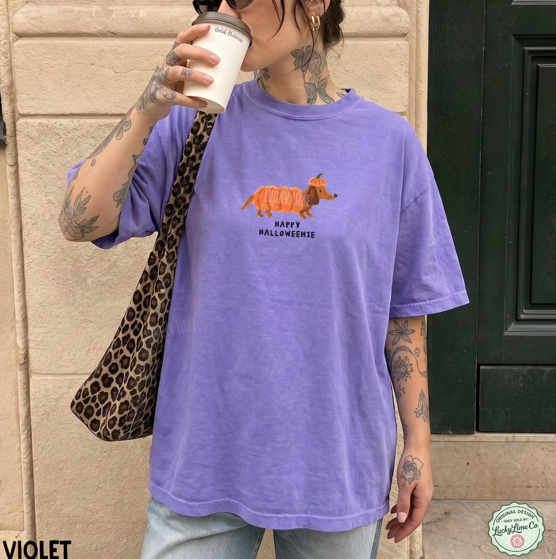 A woman wearing an oversized violet t-shirt with a pumpkin-shaped fox graphic and 'Happy Halloween' text, holding a coffee cup and wearing a leopard print bag against a beige stone wall.