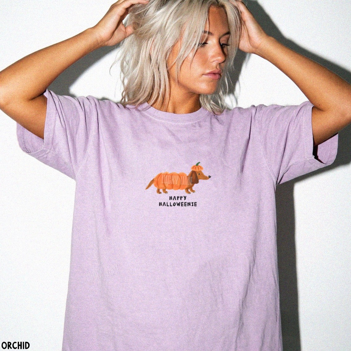 A woman with platinum blonde hair poses against a white wall wearing a lavender t-shirt with a cartoon dachshund in a pumpkin costume and the text 'HAPPY HALLOWEENIE'.