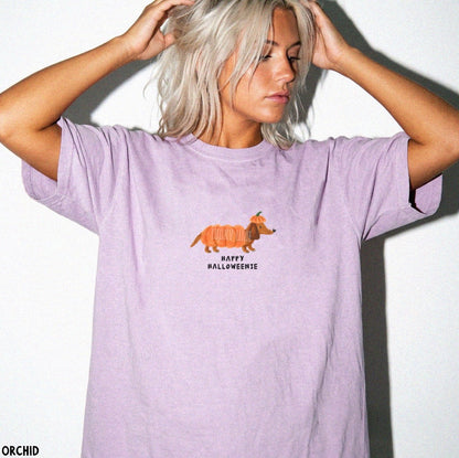 A woman with platinum blonde hair poses against a white wall wearing a lavender t-shirt with a cartoon dachshund in a pumpkin costume and the text 'HAPPY HALLOWEENIE'.