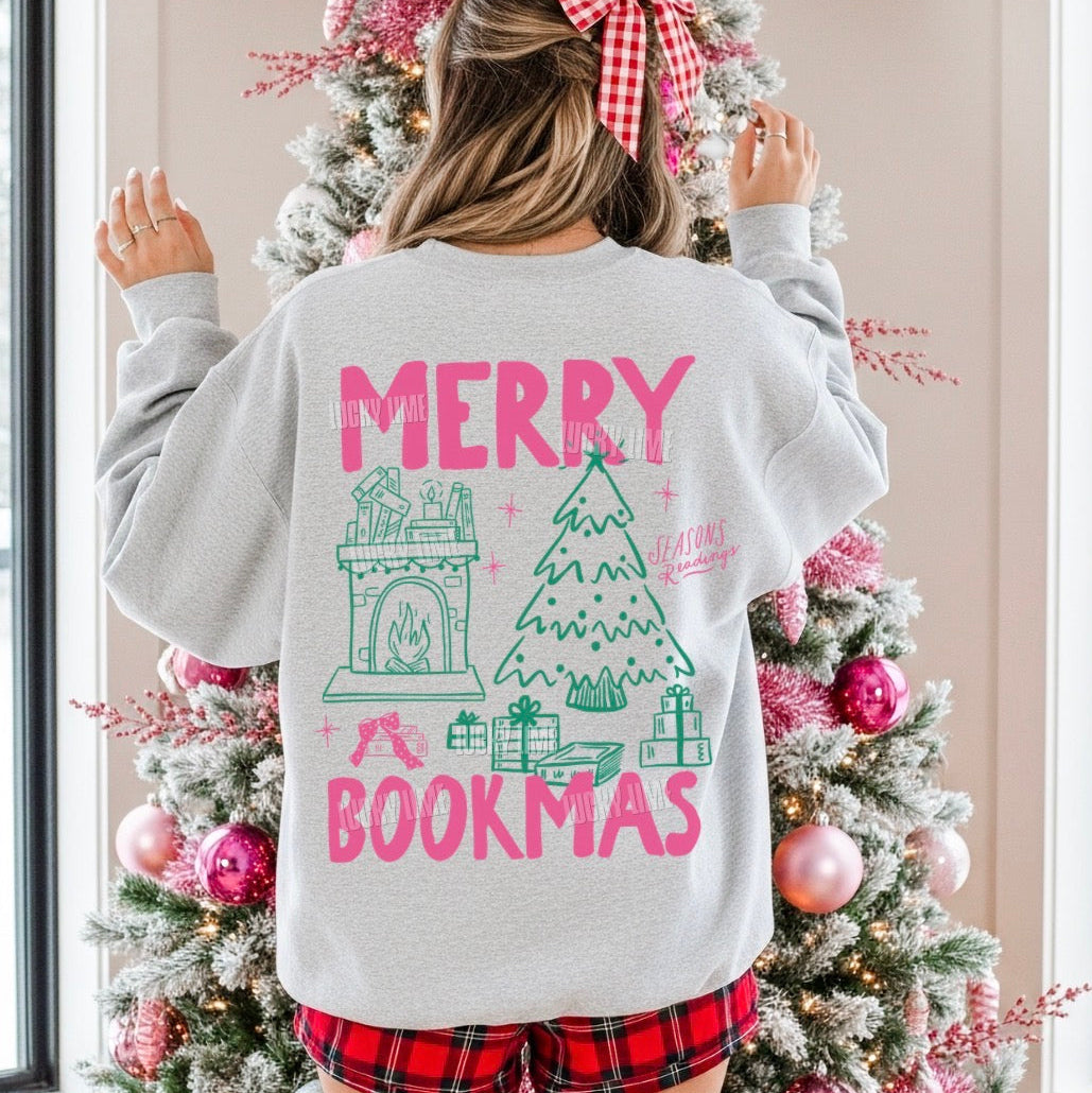 A woman viewed from behind wearing a light gray crewneck sweatshirt with a colorful Christmas-themed book-themed graphic on the back. The graphic features pink text reading 'MERRY BOOKMAS' and green illustrations of a fireplace, a Christmas tree, and wrapped presents.