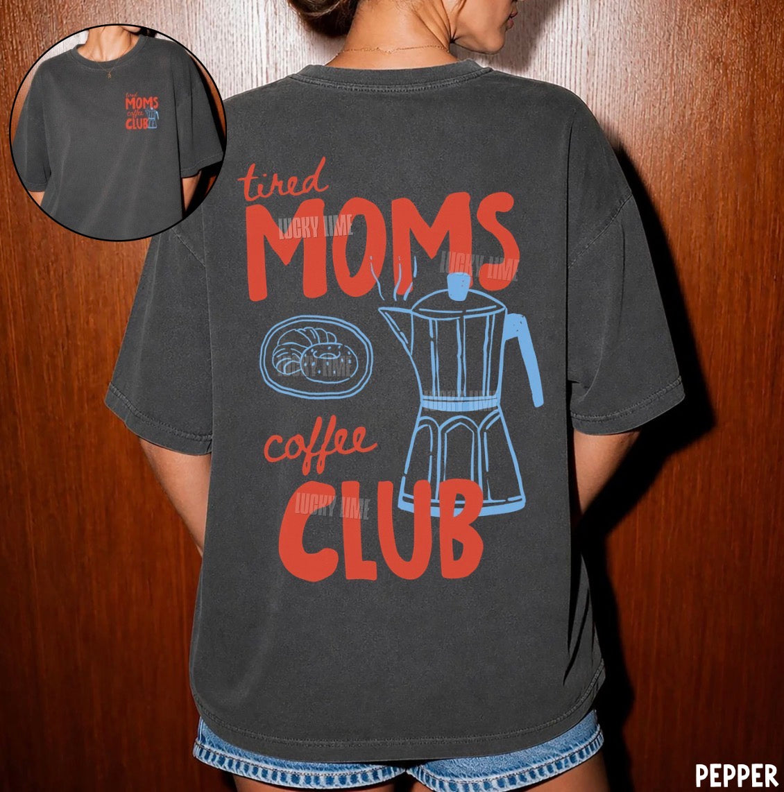 A person wearing a dark charcoal gray graphic t-shirt with a retro design that reads 'tired MOMS coffee CLUB' in red and light blue, featuring an illustration of a moka pot and croissant. A circular inset in the top-left shows the front of the t-shirt with a small red 'MOMS COFFEE CLUB' logo.