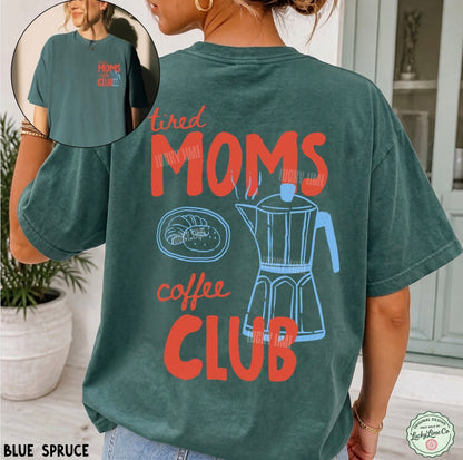 A woman wearing a faded blue-green graphic t-shirt with the text 'tired MOMS coffee CLUB' and illustrations of a plate of pastries and a moka pot. The image features a circular inset showing the front of the t-shirt with the text 'Home Club'.