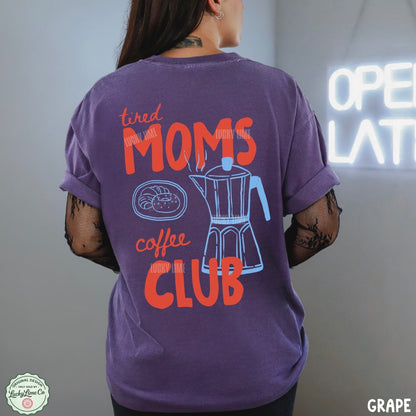 A woman shown from the back wearing a purple graphic t-shirt with the text 'tired MOMS coffee CLUB' in orange and a blue coffee maker illustration. The background is a gray wall with a glowing neon sign.