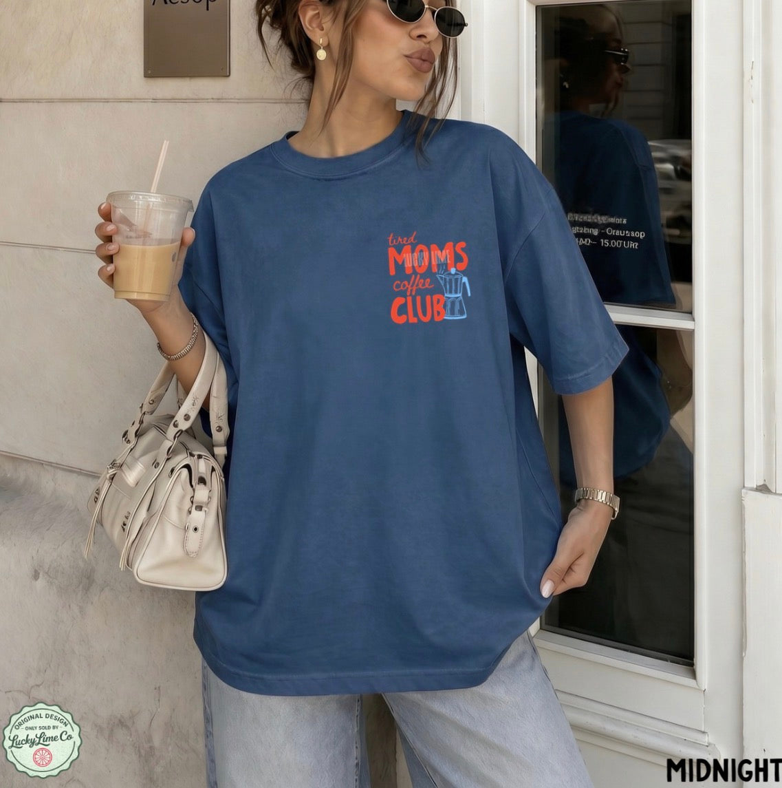 A woman wearing an oversized slate blue graphic t-shirt with the text 'tired MOMS coffee CLUB' in red and orange, paired with light-wash jeans and holding a coffee cup.