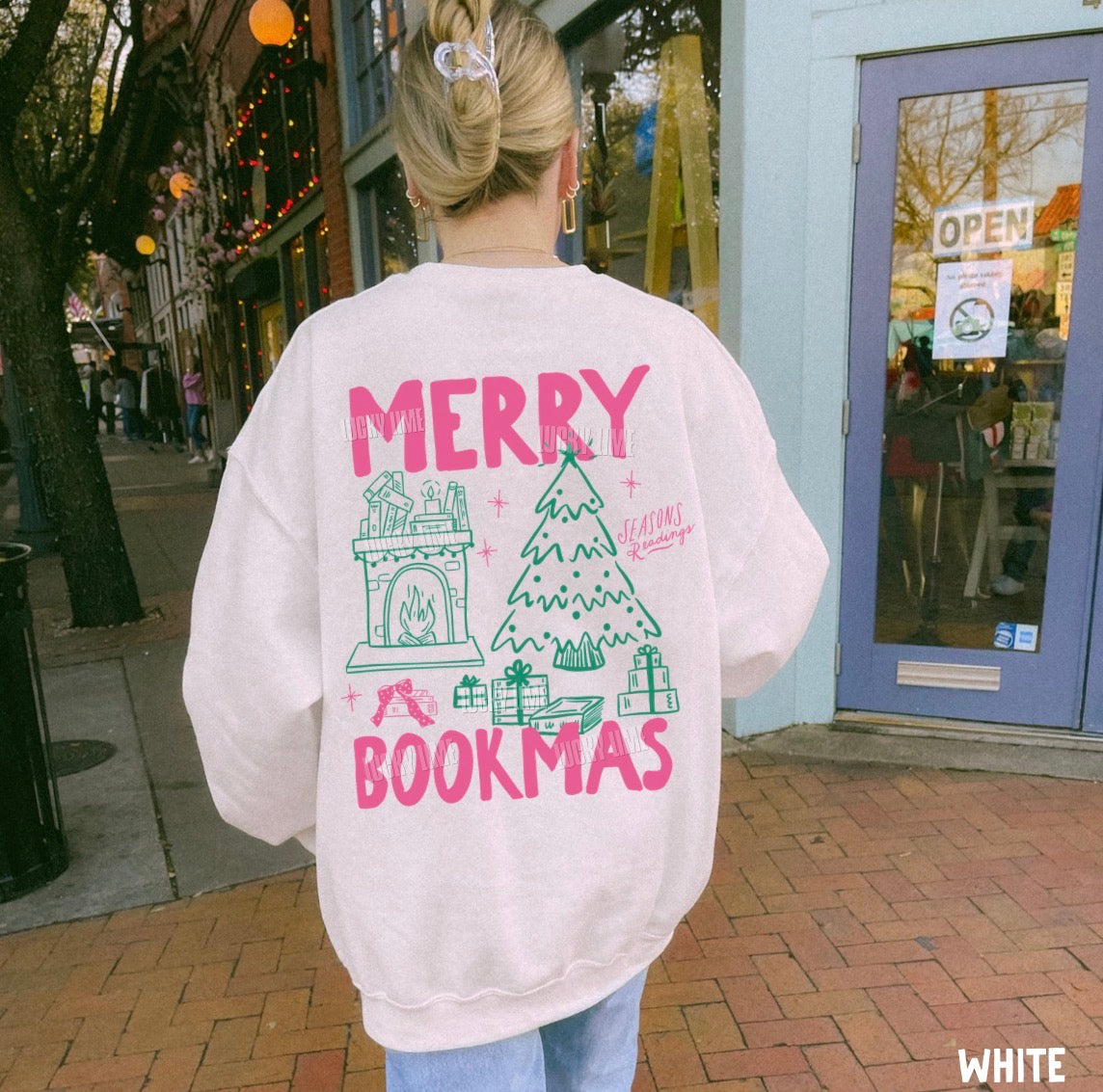 A woman viewed from behind wearing a light pink sweatshirt with a large 'MERRY BOOKMAS' graphic in hot pink and green on the back, standing on a brick sidewalk in front of a storefront.