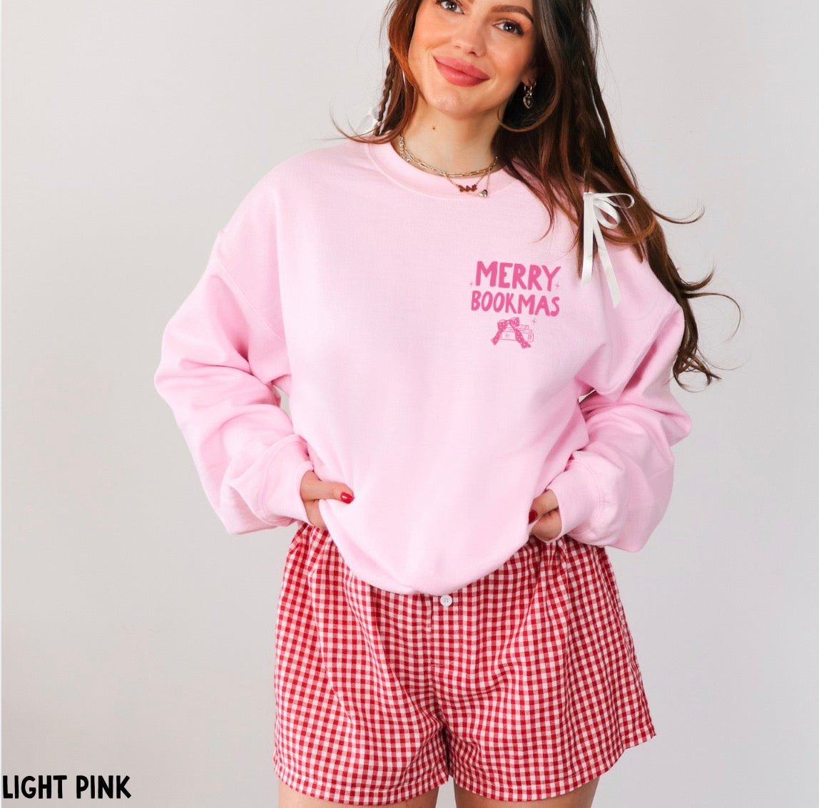 A woman wearing a light pink sweatshirt with 'MERRY BOOKMAS' text on the chest and red-and-white gingham shorts, posed against a light gray studio background.