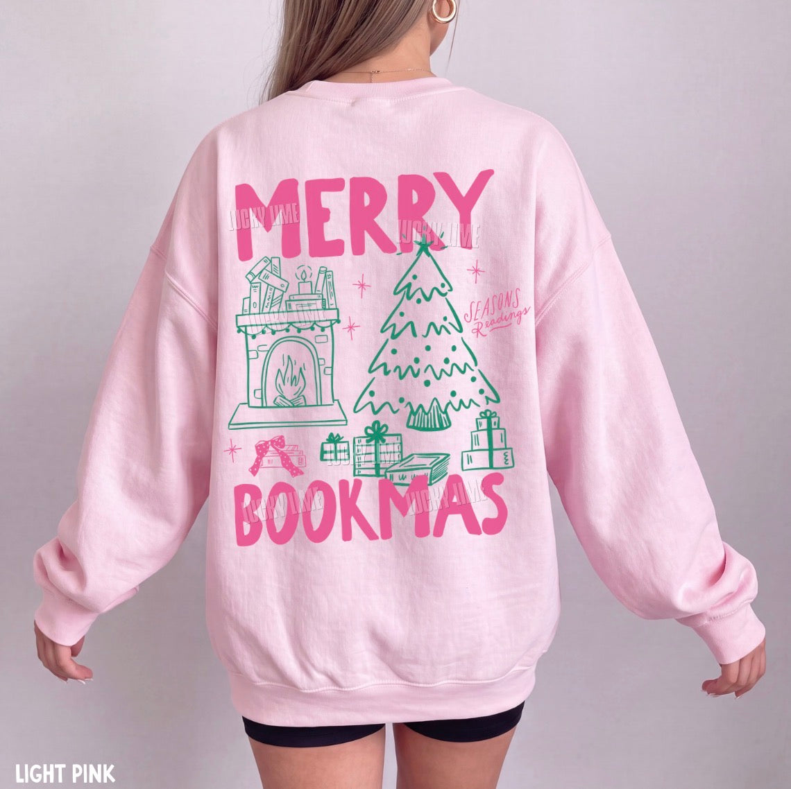 A person shown from the back wearing a light pink crewneck sweatshirt with a large 'MERRY BOOKMAS' graphic featuring a fireplace, Christmas tree, and gifts in pink and green. The image has a text overlay 'LIGHT PINK' in the bottom-left corner.