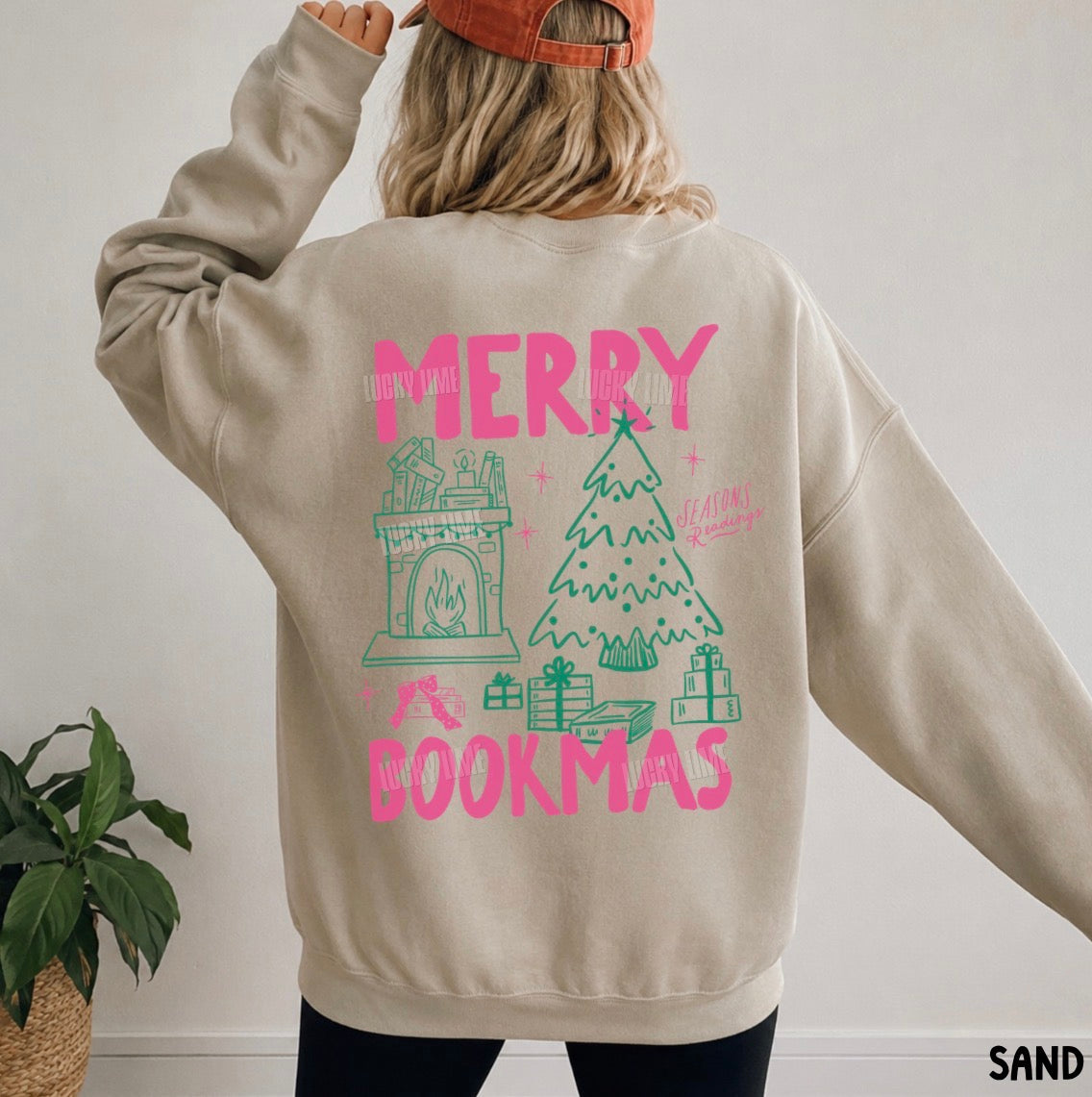 A person wearing a beige crewneck sweatshirt with a colorful graphic on the back that reads 'MERRY BOOKMAS' in pink, featuring illustrations of a fireplace, Christmas tree, and wrapped gifts. The person is also wearing an orange cap and black pants, standing indoors next to a potted plant.