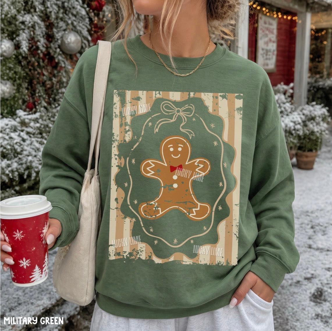 A woman wearing a military green crewneck sweatshirt with a vintage-style gingerbread man graphic on the front, holding a red holiday coffee cup in a snowy outdoor setting.