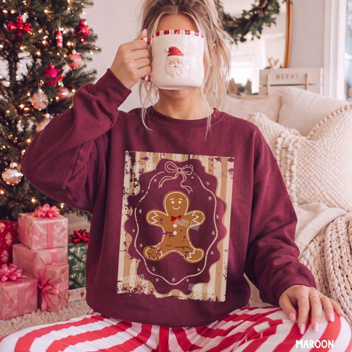 A woman in a cozy living room setting wearing a maroon crewneck sweatshirt with a gingerbread man graphic, holding a white mug with a Santa face. A decorated Christmas tree and wrapped gifts are visible in the background.