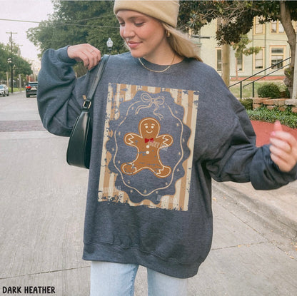 A woman wearing an oversized dark heather gray sweatshirt with a large gingerbread man graphic on the front, paired with a tan beanie and light blue jeans, standing on a city street.