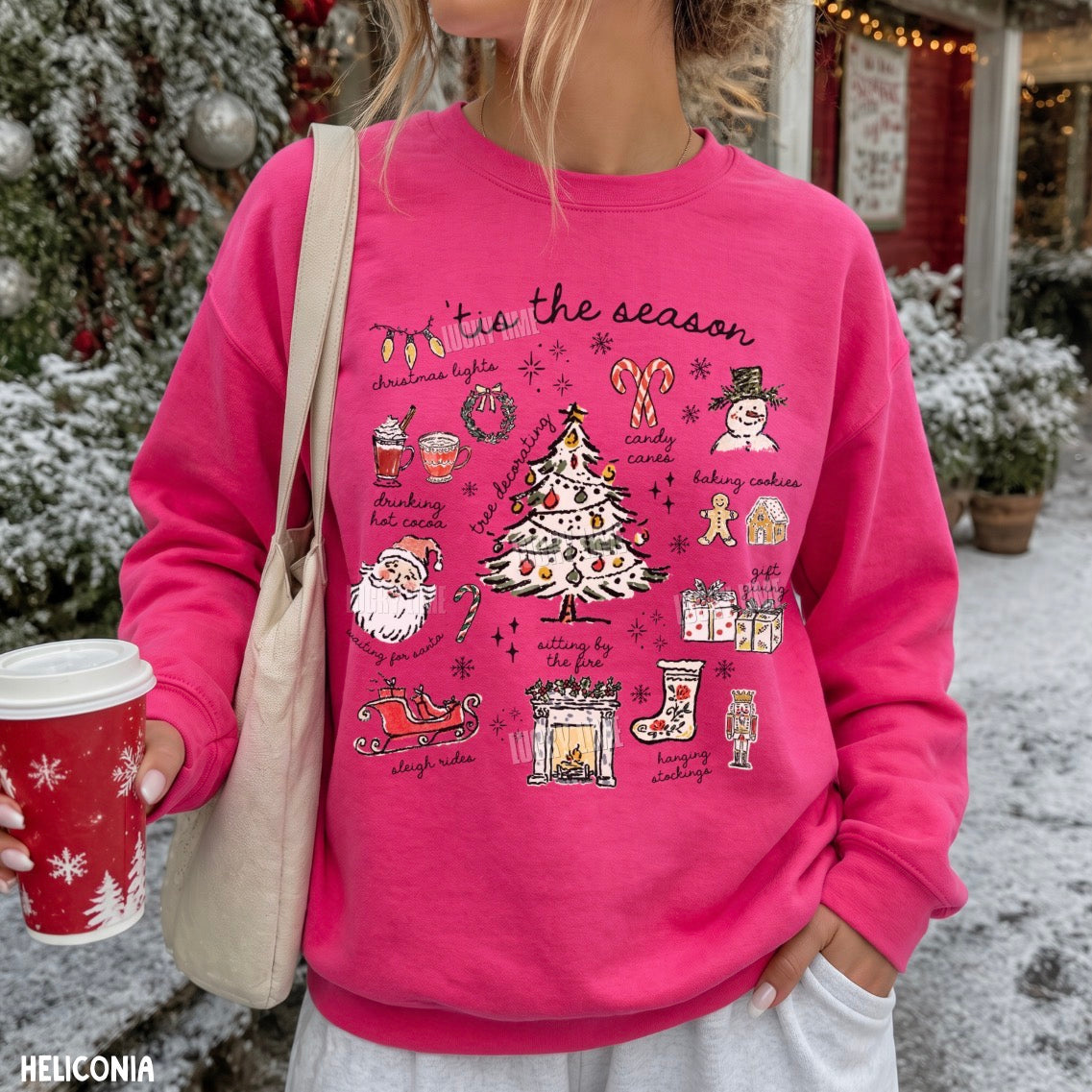 A woman wearing a bright pink crewneck sweatshirt with a detailed Christmas graphic that reads 'Tie the season' and features illustrations of a decorated tree, Santa, candy canes, gingerbread cookies, and stockings. She is holding a red holiday coffee cup and wearing a beige shoulder bag, set against a snowy outdoor background with Christmas decorations.