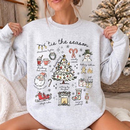 A woman wearing a light heather gray crewneck sweatshirt with a detailed Christmas-themed graphic print that reads 'Tis the season' and includes illustrations of a Christmas tree, Santa, stockings, candy canes, and a sleigh, set in a cozy holiday home environment.