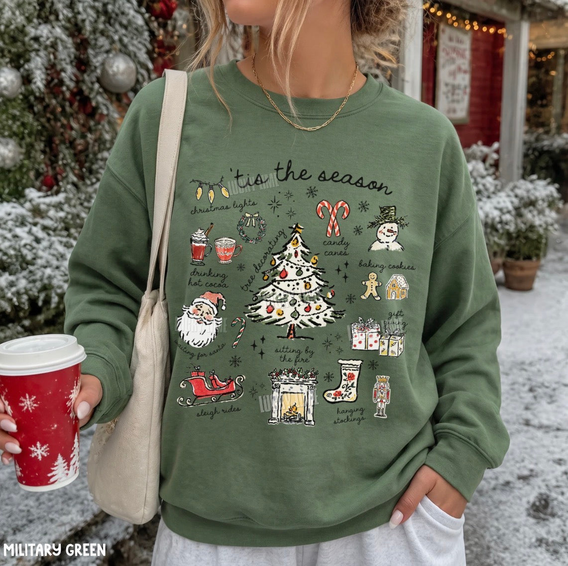 A woman wearing a military green crewneck sweatshirt with a colorful Christmas graphic print that reads 'tie the season' and features illustrations of a Christmas tree, Santa Claus, candy canes, and snowman. She is holding a red holiday coffee cup and a beige shoulder bag, standing outdoors in a snowy, decorated holiday setting.