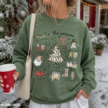 A woman wearing a military green crewneck sweatshirt with a colorful Christmas graphic print that reads 'tie the season' and features illustrations of a Christmas tree, Santa Claus, candy canes, and snowman. She is holding a red holiday coffee cup and a beige shoulder bag, standing outdoors in a snowy, decorated holiday setting.
