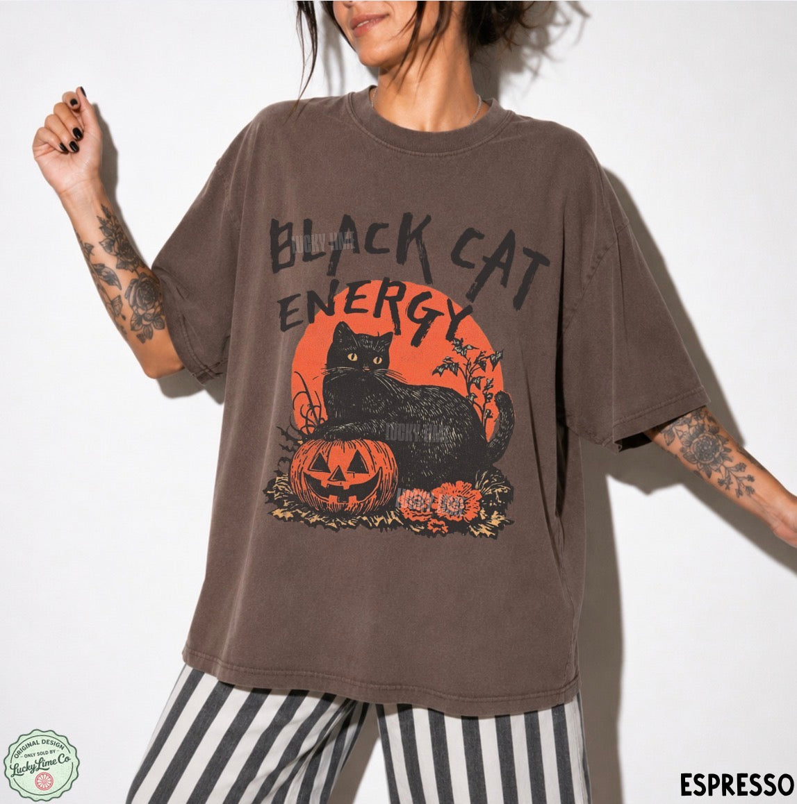 A woman wearing an oversized brown graphic t-shirt with a black cat and jack-o'-lantern design, paired with black and white striped pants, against a white background.