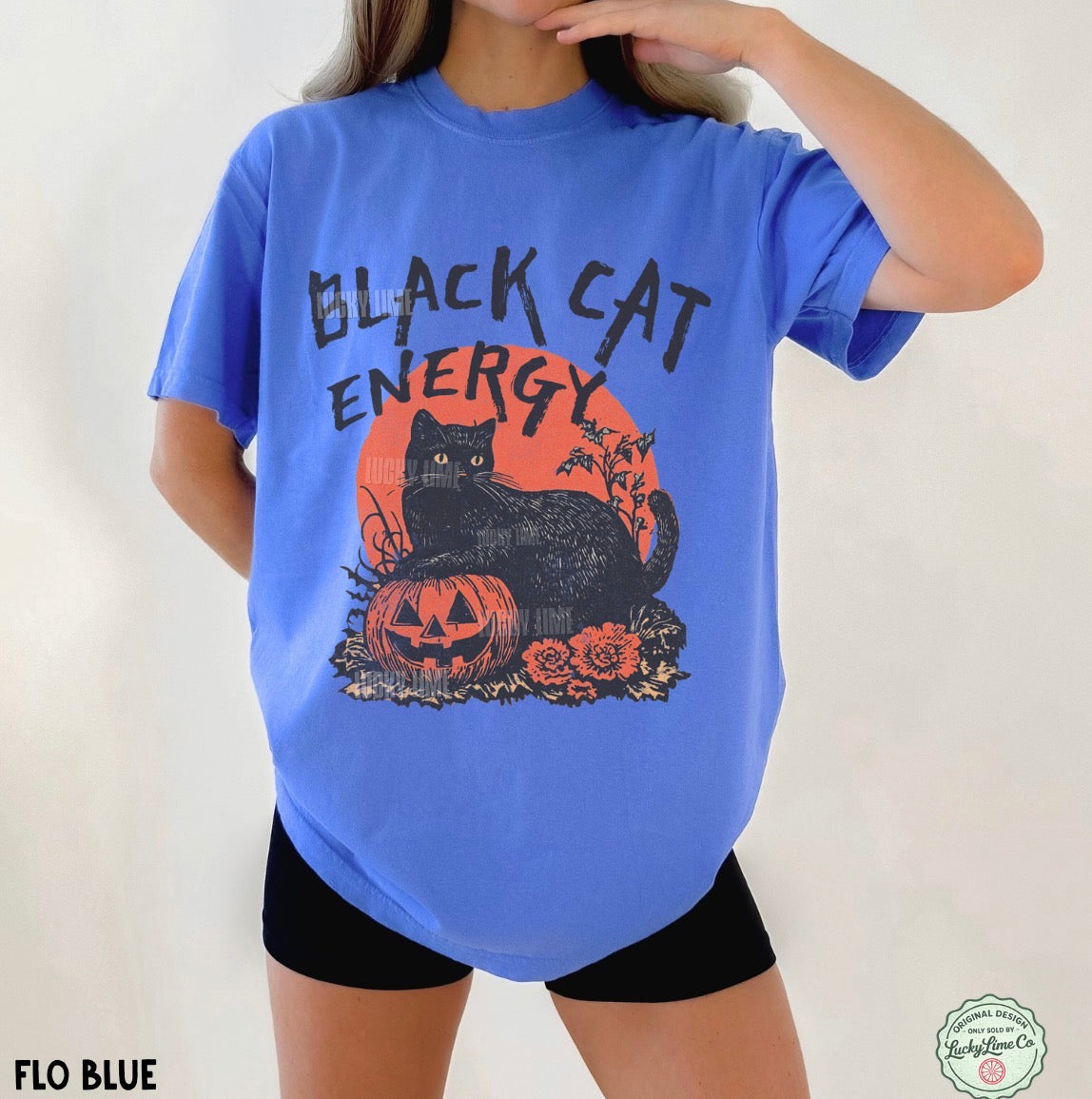 A person wearing an oversized bright blue graphic T-shirt with a black cat, jack-o'-lantern, roses, and the words 'BLACK CAT ENERGY' printed on the front, paired with black shorts against a plain light background.