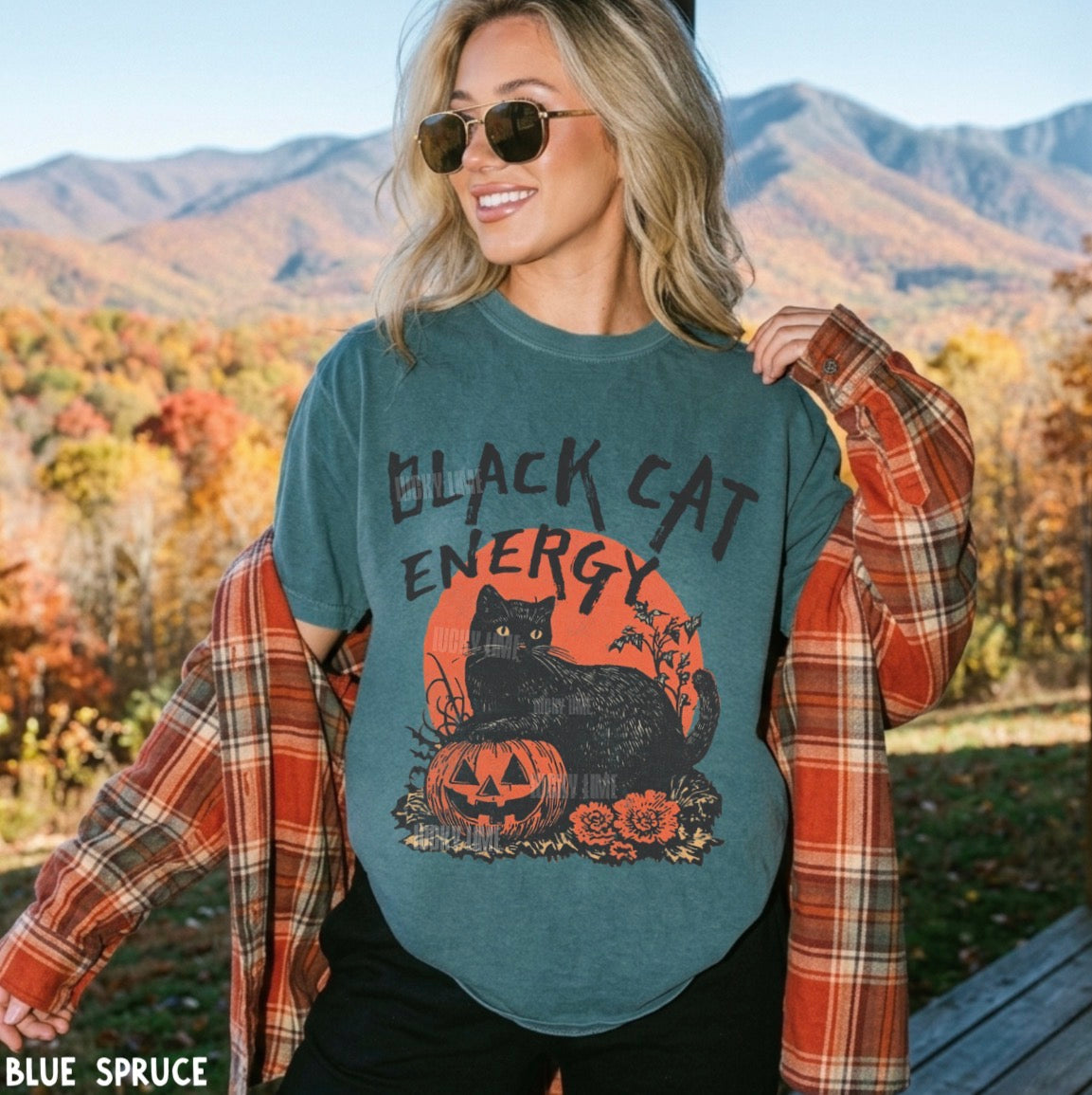 A woman wearing a slate blue graphic t-shirt with a 'Black Cat Energy' Halloween design, styled with an orange plaid flannel shirt and sunglasses, set against a scenic autumn mountain background.