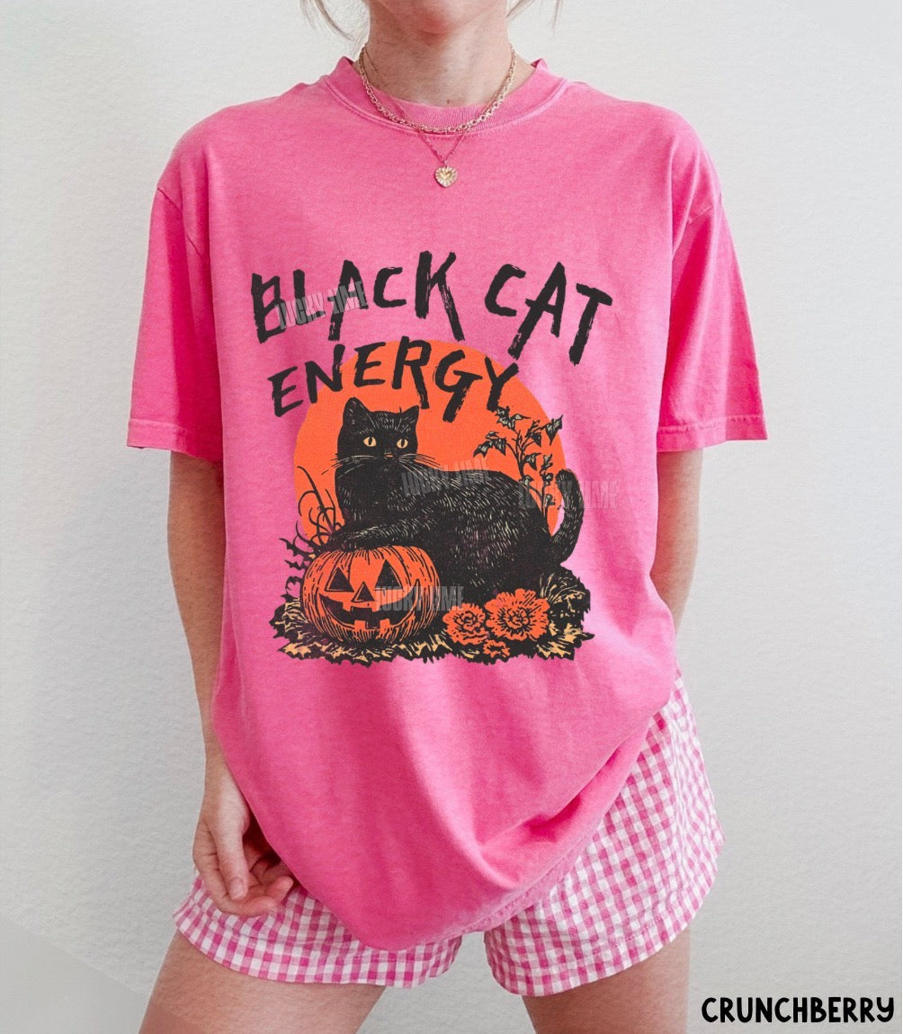 A person wearing a bright pink graphic T-shirt with a black cat, orange sun, and jack-o'-lantern design reading 'BLACK CAT ENERGY,' paired with pink-and-white gingham shorts against a plain light wall.