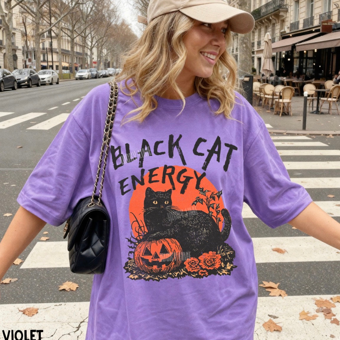 A woman wearing a purple oversized graphic T-shirt with a black cat and orange pumpkin design, standing on a city crosswalk. The shirt reads 'BLACK CAT ENERGY' above a black cat over a jack-o'-lantern and orange circle.