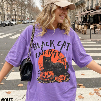 A woman wearing a purple oversized graphic T-shirt with a black cat and orange pumpkin design, standing on a city crosswalk. The shirt reads 'BLACK CAT ENERGY' above a black cat over a jack-o'-lantern and orange circle.