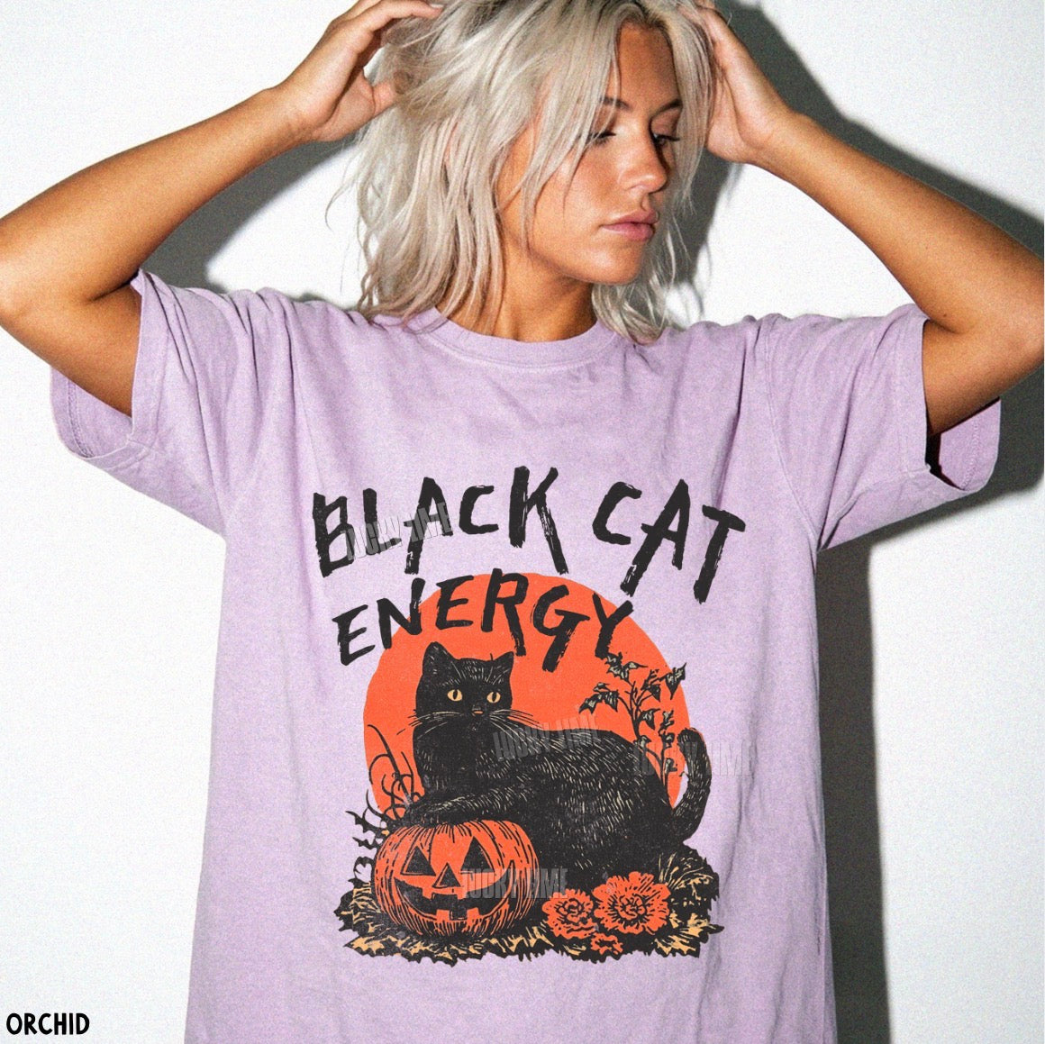 A woman wearing an oversized light purple t-shirt with a graphic of a black cat, a jack-o'-lantern, and an orange sun with the text 'BLACK CAT ENERGY'.
