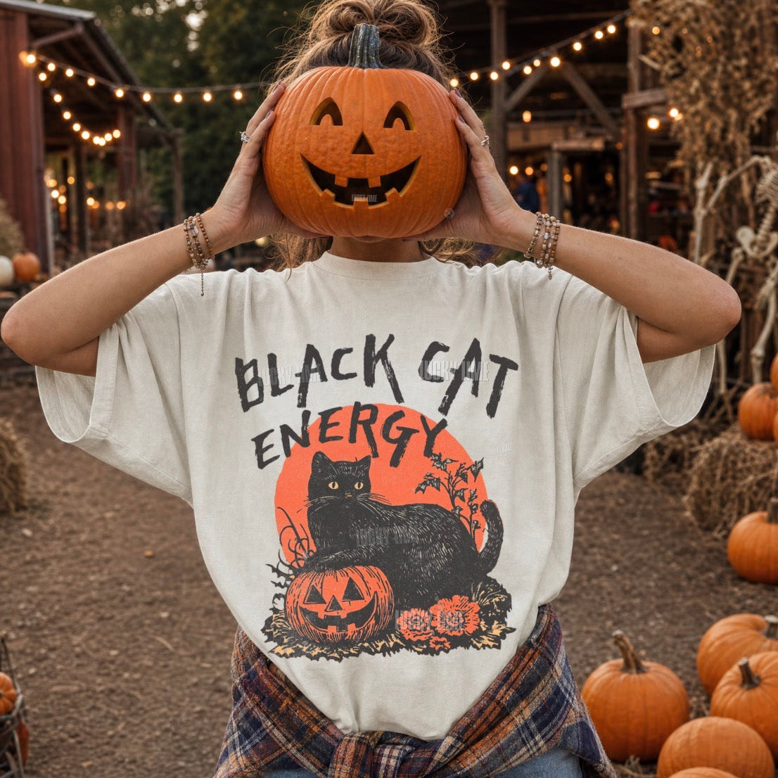 A person outdoors at a pumpkin patch wearing an oversized cream graphic T-shirt with 'BLACK CAT ENERGY' text and a black cat illustration over an orange circle, holding a carved pumpkin to their face.