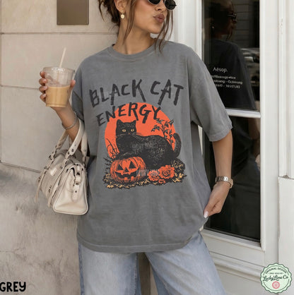 A woman wearing an oversized grey graphic t-shirt with a 'Black Cat Energy' Halloween print, paired with light wash jeans and holding a drink and a handbag.