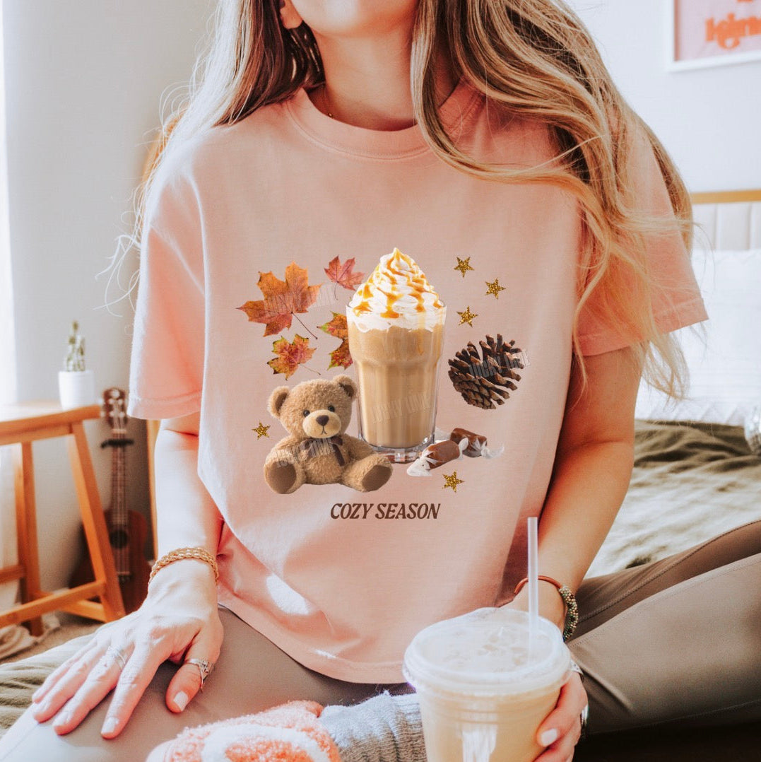 A woman wearing a peach-pink graphic t-shirt in a cozy bedroom setting. The t-shirt features a 'Cozy Season' graphic with a teddy bear, a hot coffee drink, autumn leaves, and a pinecone.