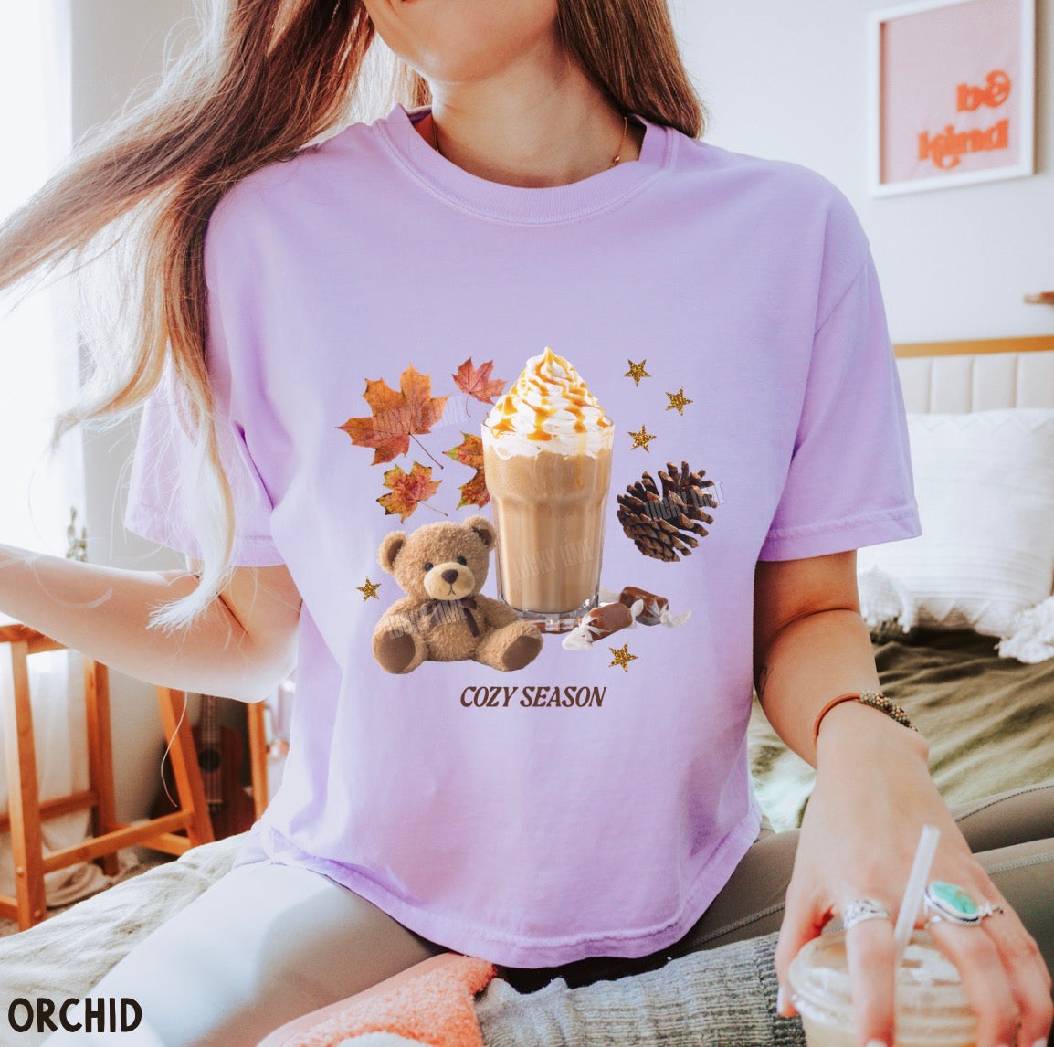 A woman wearing a lavender graphic t-shirt with a 'Cozy Season' design featuring a hot cocoa drink, a teddy bear, autumn leaves, and a pinecone. The brand name 'ORCHID' is visible in the bottom-left corner.