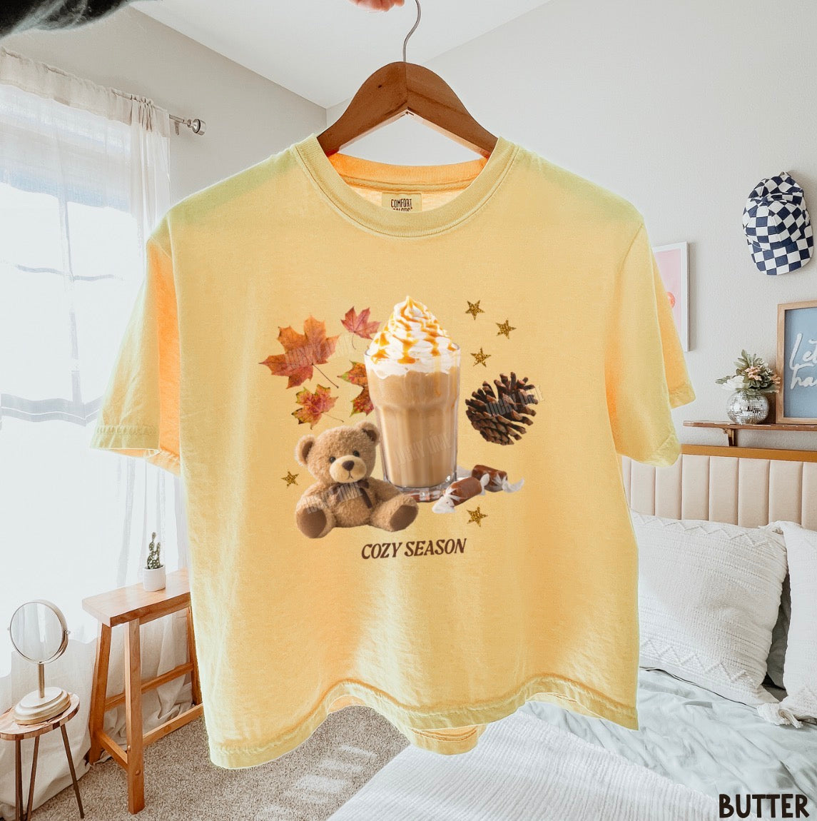 A pale yellow short-sleeve graphic T-shirt hanging on a wooden hanger in a bedroom setting. The shirt features a cozy autumn-themed graphic with a teddy bear, a hot coffee drink, maple leaves, pinecones, and the text 'COZY SEASON'.
