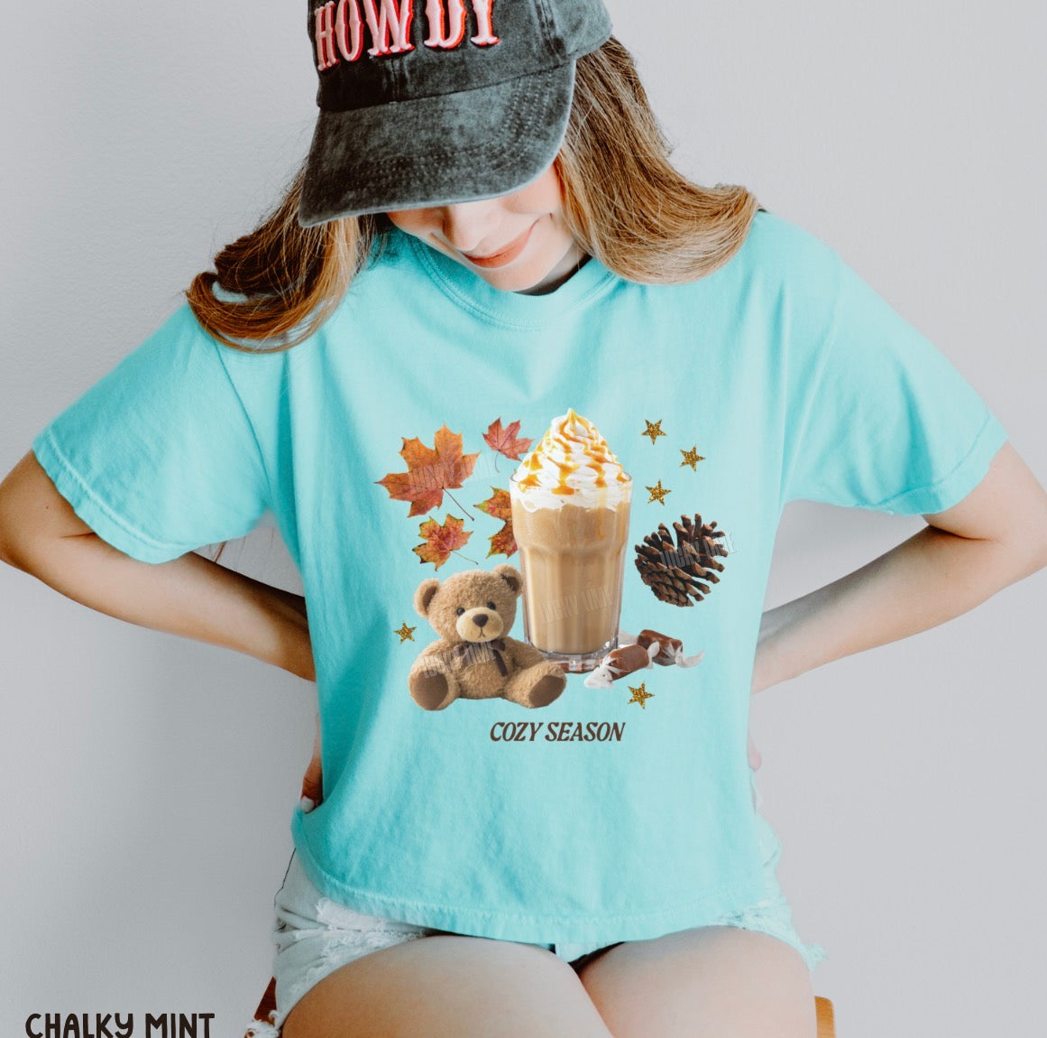 A person wearing a light aqua graphic T-shirt with a fall-themed print of a teddy bear, iced coffee, autumn leaves, and a pinecone, paired with a dark gray cap and denim shorts.