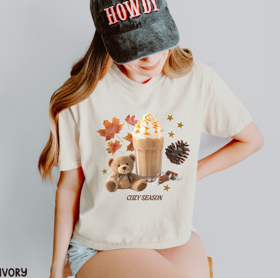 A woman wearing a cream-colored graphic t-shirt with a 'COZY SEASON' autumn theme print, paired with a black cap that says 'HOWDY'.