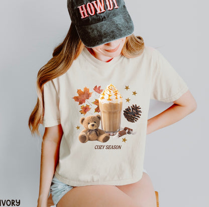 A woman wearing a cream-colored graphic t-shirt with a 'COZY SEASON' autumn theme print, paired with a black cap that says 'HOWDY'.