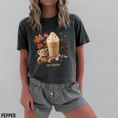 A woman wearing a dark charcoal graphic t-shirt with a cozy autumn-themed print (teddy bear, iced coffee, fall leaves) and black-and-white gingham shorts. The text 'PEPPER' is visible in the bottom-left corner.
